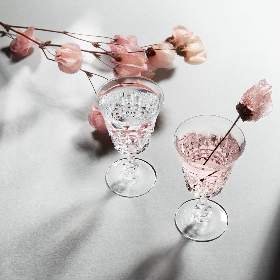 Two crystal glasses, one with clear liquid, one with pale pink liquid, accented by light-pink flowers on a reflective grey surface.