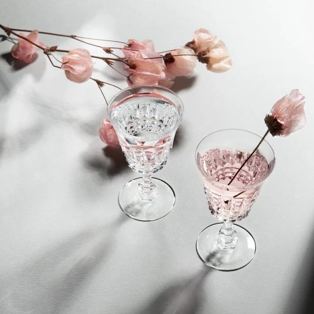 Two crystal glasses, one with clear liquid, one with pale pink liquid, accented by light-pink flowers on a reflective grey surface.