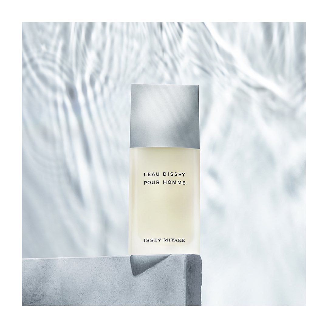 Still life of L'Eau d'Issey Pour Homme fragrance bottle by Issey Miyake on a grey concrete block against water-patterned backdrop.