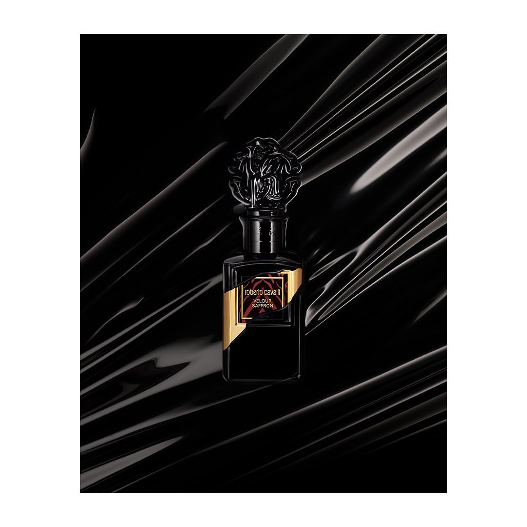 Black perfume bottle with ornate cap on glossy dark draped fabric background photographed by Alizee Patton