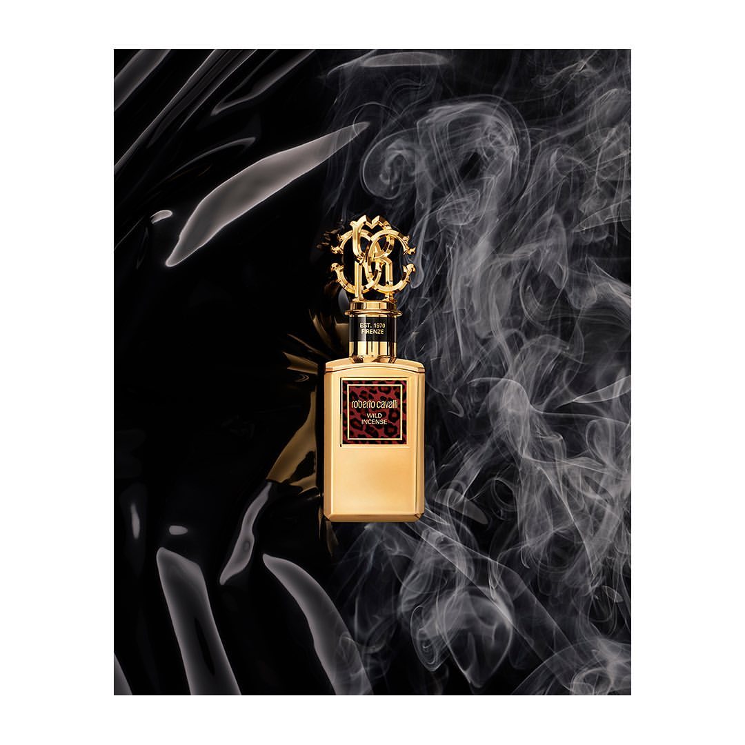Gold Roberto Cavalli Wild Laces perfume bottle with elaborate gold topper on a black surface with smoke. Beauty and fragrance ad.
