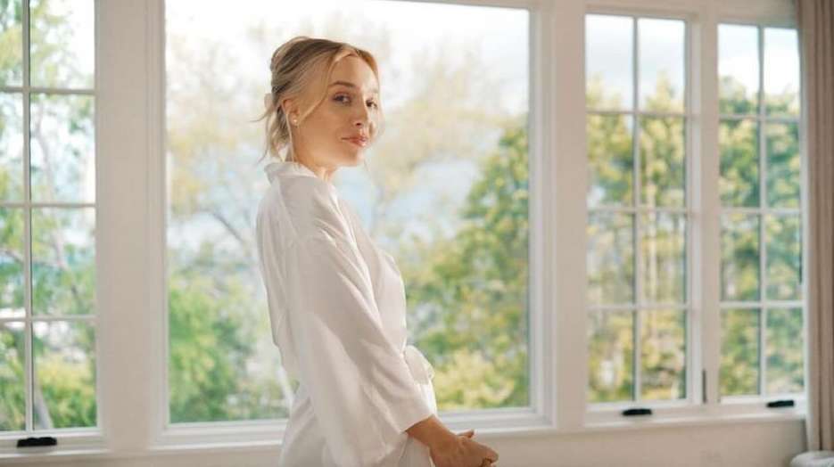 Woman in white robe poses by a large window overlooking trees and greenery in an indoor setting with soft, natural light.