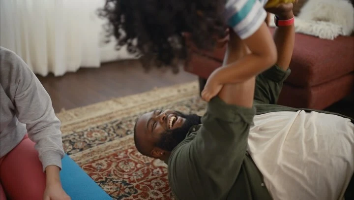 A happy Black father lies on a rug, holding his child above him with a smiling woman beside a yoga mat in an indoor setting.