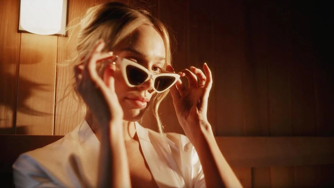 Blonde woman poses indoors wearing a white robe and white sunglasses, gazing slightly off-camera with a subtle smile.