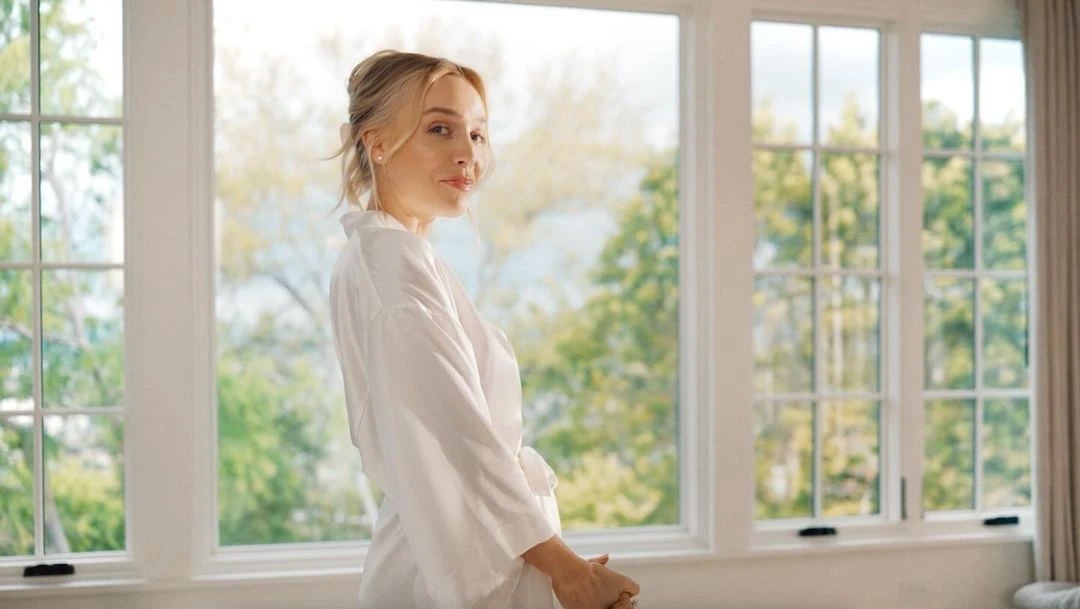 Woman in white robe looks back from window inside a light-filled home with a view of trees, shot in soft focus during daylight.