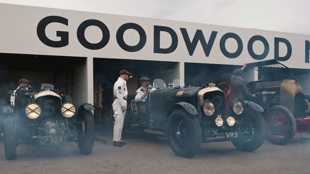 Vintage race cars on display at Goodwood Motor Circuit with drivers in vintage racing suits and "GOODWOOD" signage.