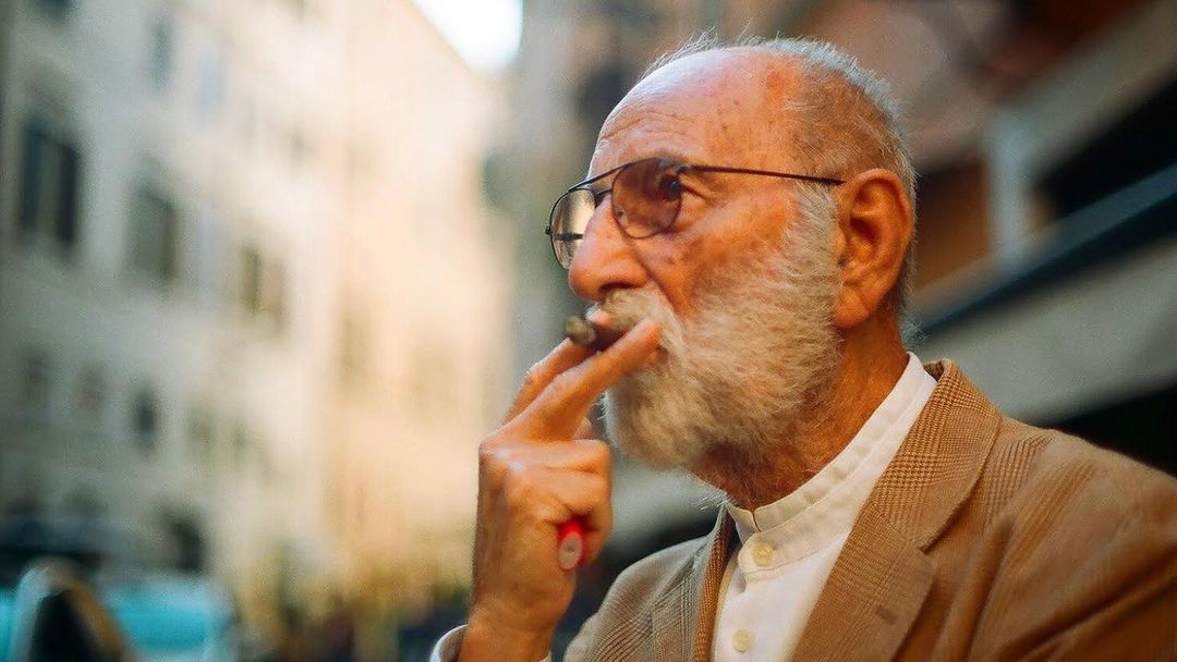 Elderly man with white beard and glasses in a tan jacket smoking a cigar outdoors against an out-of-focus urban backdrop.