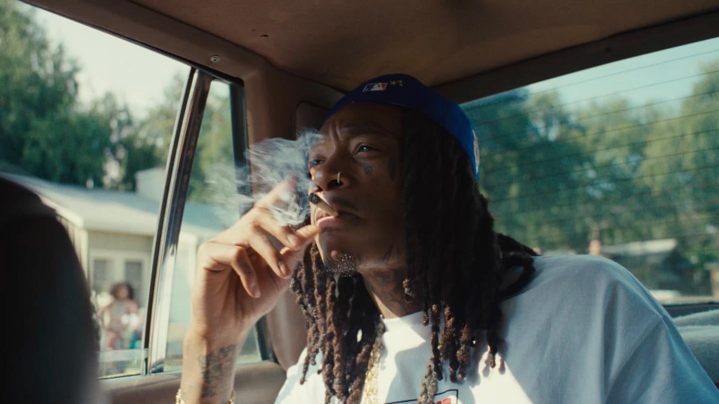 Portrait of adult male in a car with a blue baseball cap smoking, with trees and a house visible through the car window.