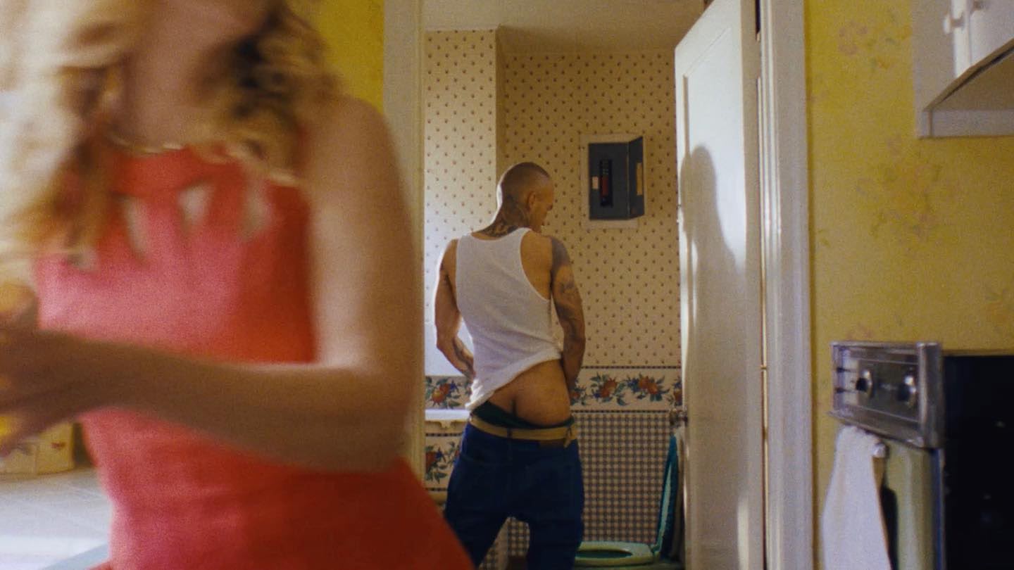 Man with tattoos pulling down his pants in a yellow bathroom with a woman in a pink dress partially visible in the foreground.
