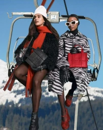 Two women seated on a ski lift chair holding branded tote bags against a snowy mountain backdrop.