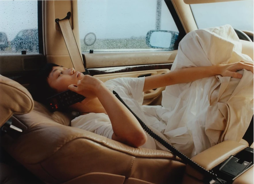 An Asian woman reclines in the tan leather interior of a car while talking on a mobile phone in front of a rainy window.