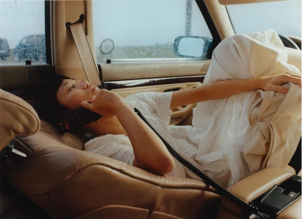 An Asian woman reclines in the tan leather interior of a car while talking on a mobile phone in front of a rainy window.