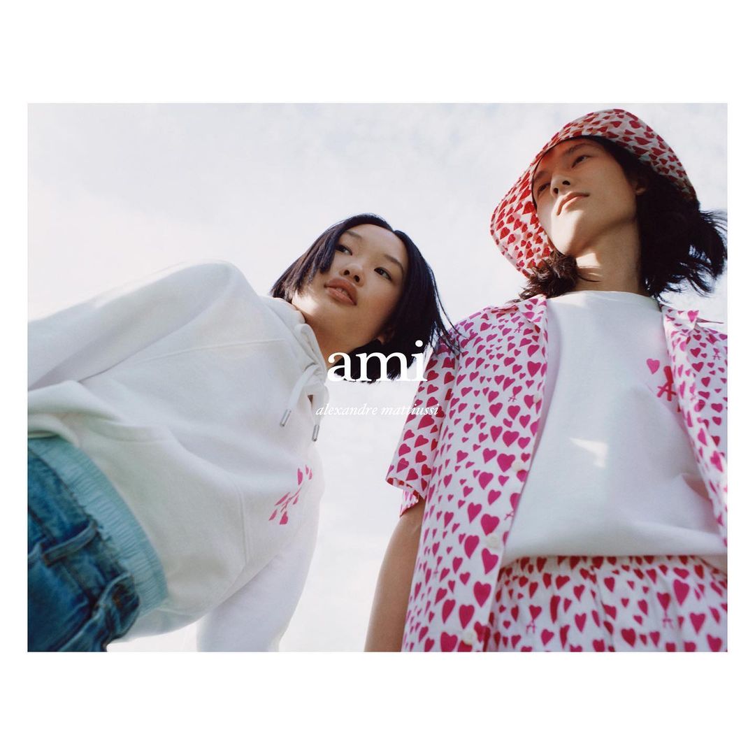 Two Asian fashion models in a low angle shot. One is wearing a heart patterned bucket hat. Branded apparel is visible against a bright sky.