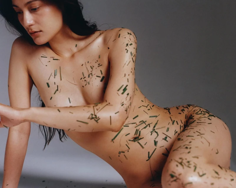 Nude woman poses in a studio with short strands of green plant material scattered on her body and lying on a grey backdrop.