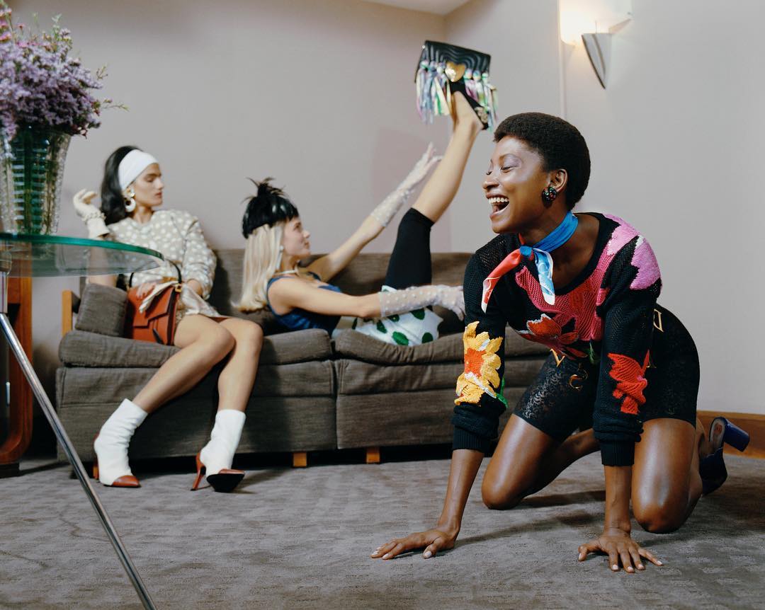 Three women pose in an indoor setting. One Black woman kneels, laughing, wearing colorful top, while the other two sit on a couch.