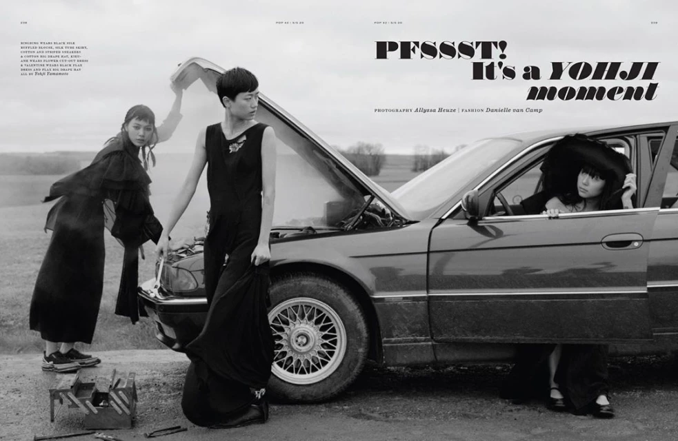 Three women in black dresses stand by a vintage car with its hood open in a rural setting. Fashion editorial photography.