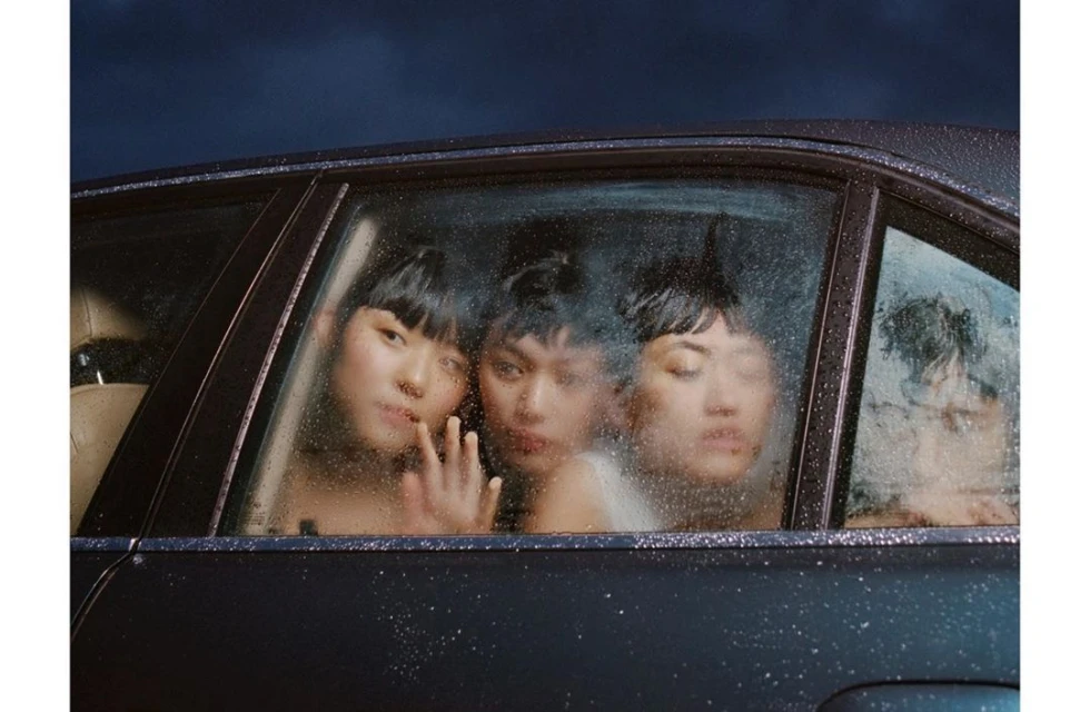 Three women with short dark hair are seen through a wet car window, reflecting soft light with a dark sky in the background.