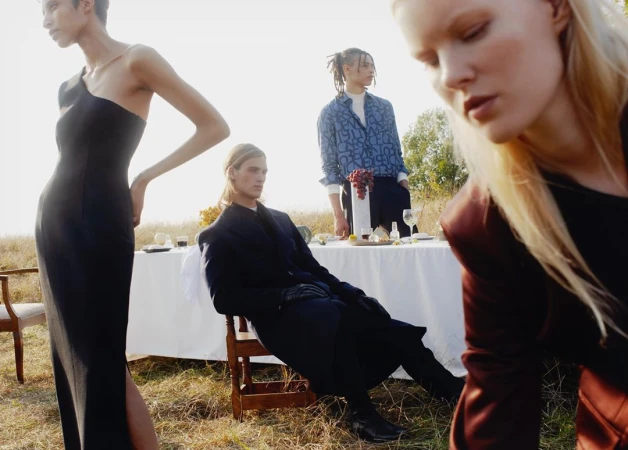Three fashion models pose outdoors at a table set with food and wine in a sunlit field, creating a sophisticated outdoor gathering scene.