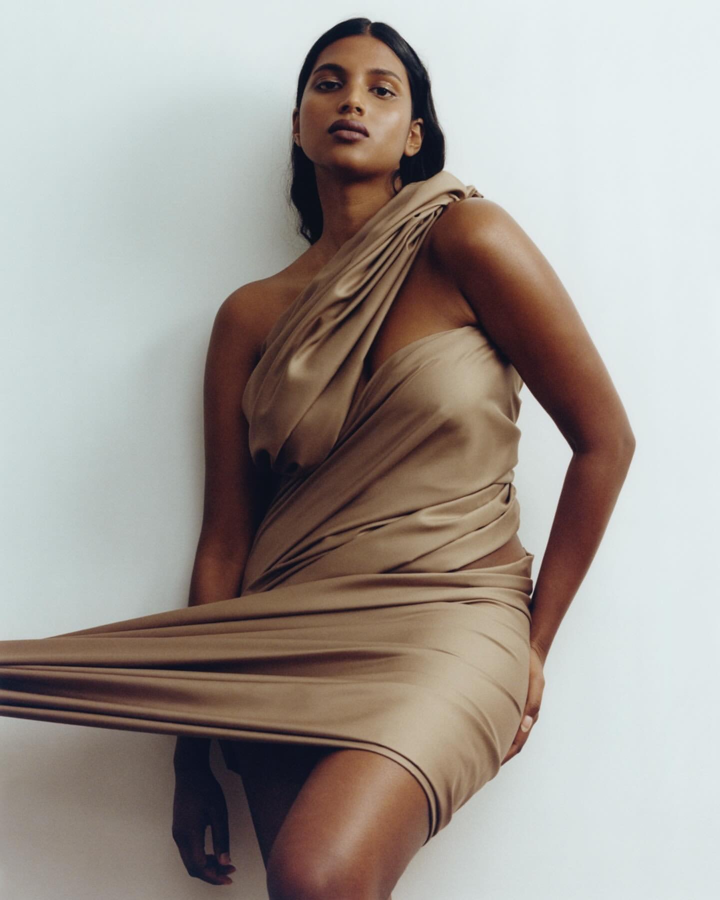 Woman with dark hair poses in a draped gold dress against a white background in a beauty shot. The image has a fashion editorial style.