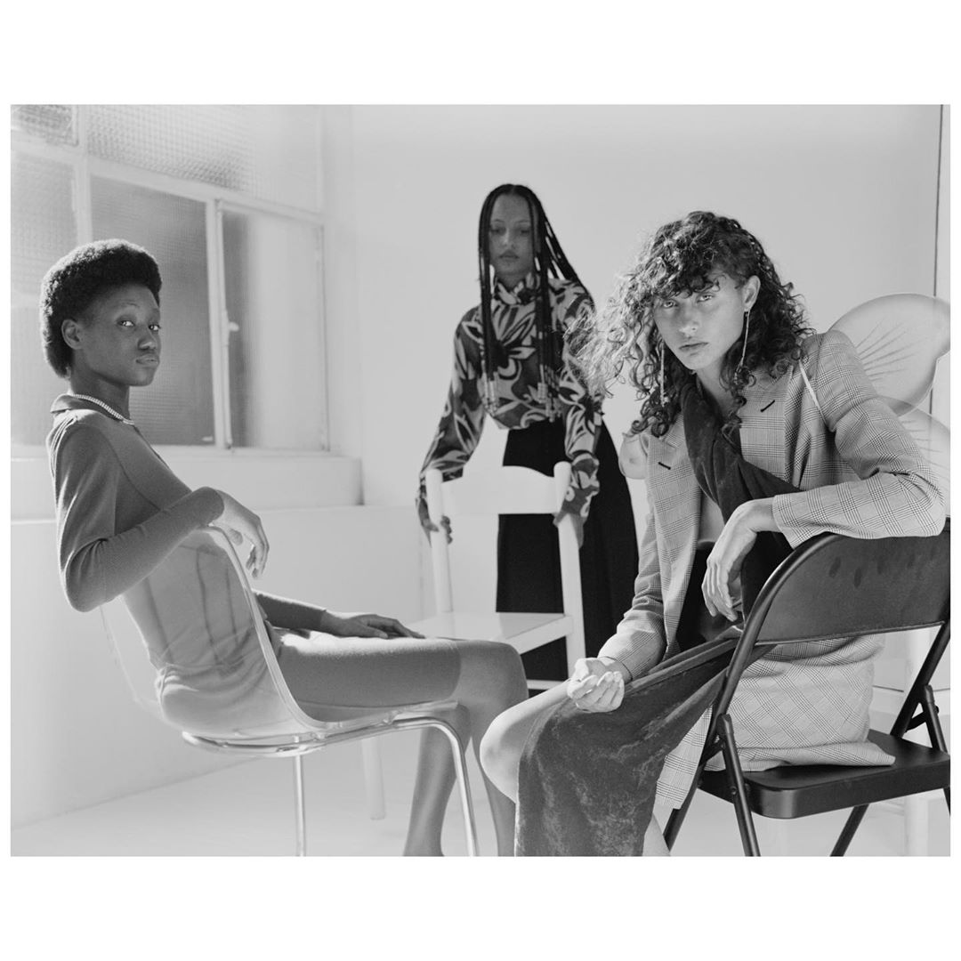 Three young adult women in fashionable clothing sit and stand in a bright studio, posing with chairs, rendered in black and white.