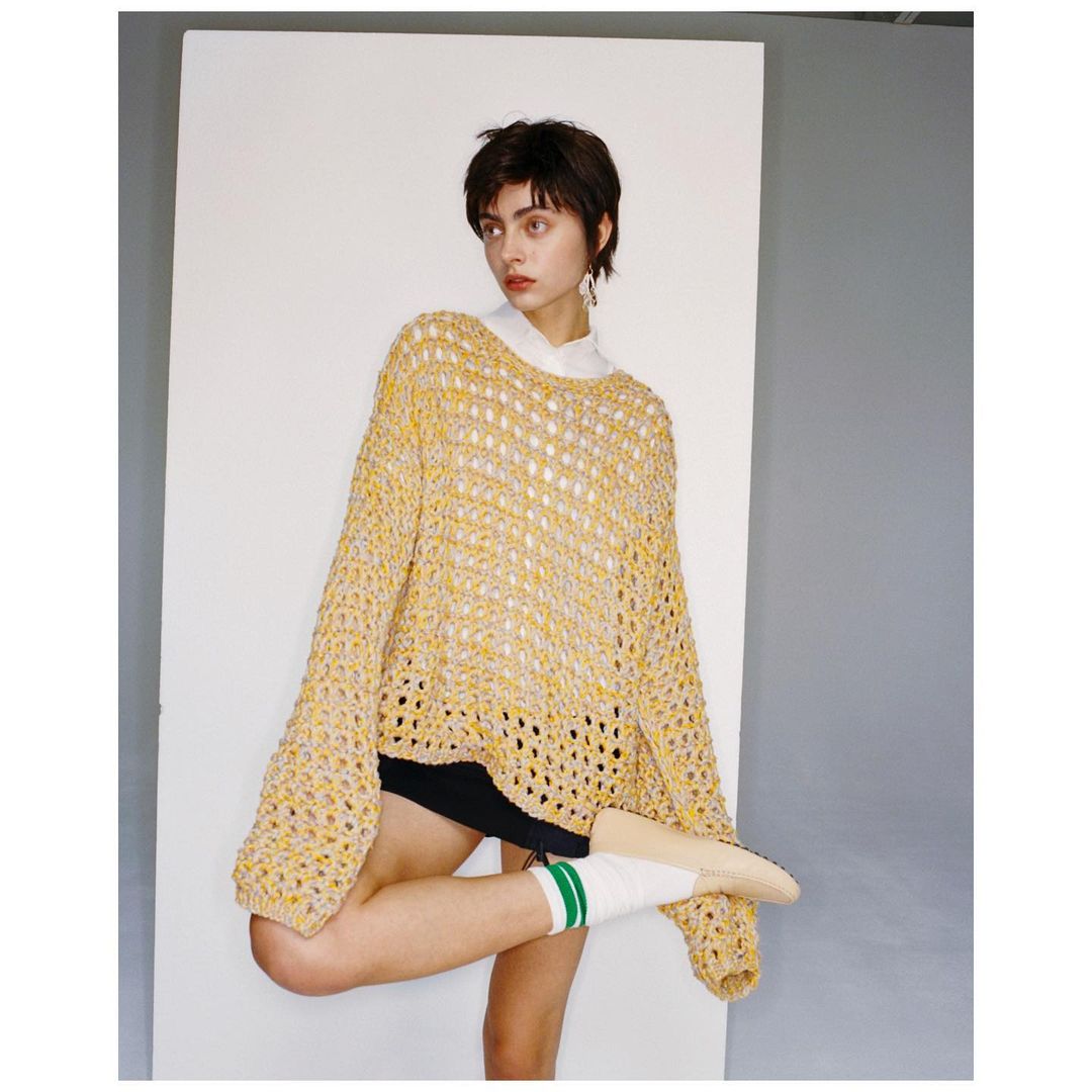 A woman poses indoors wearing a yellow, open-weave sweater, black shorts, white socks with green stripes, and beige shoes, against a neutral backdrop.
