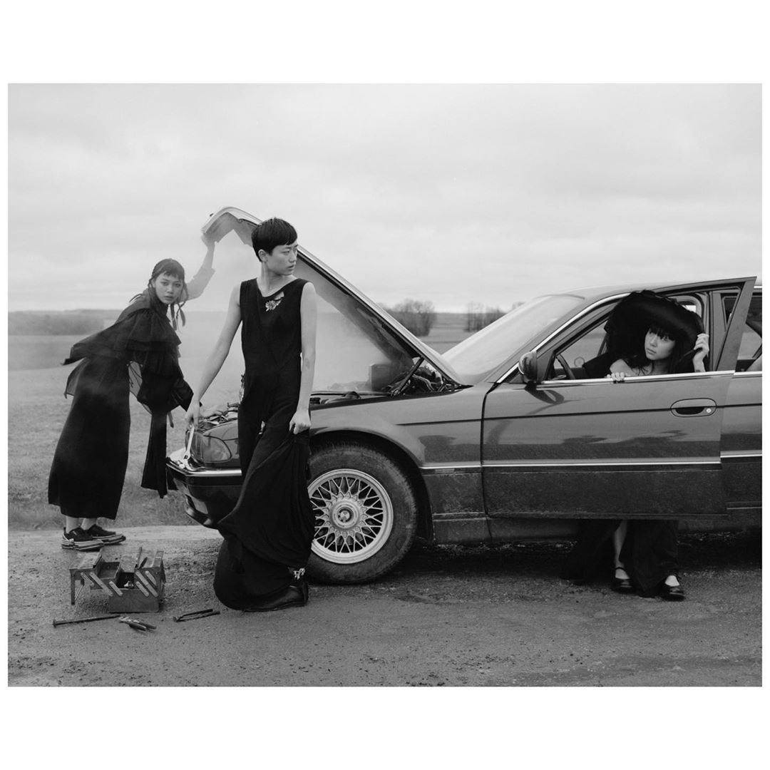 Monochromatic shot of three women in formal dresses with a broken-down car in a rural setting under an overcast sky, cinematic and editorial.