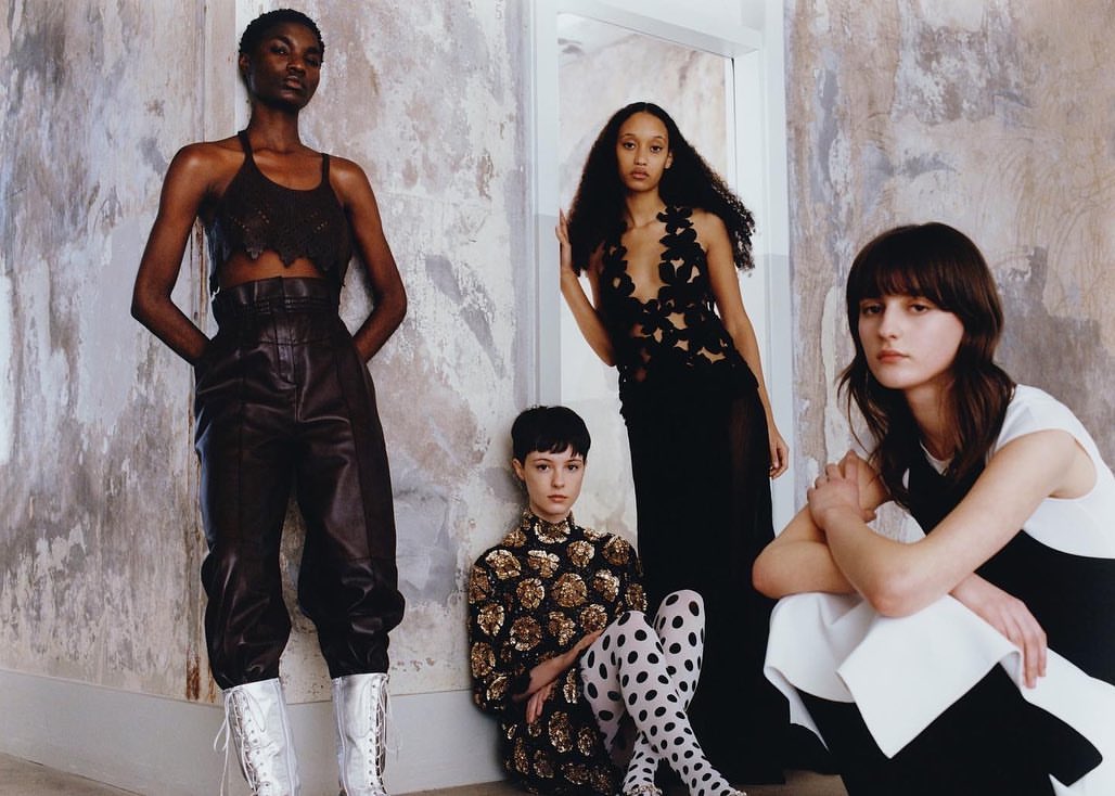Four women pose indoors wearing black and white fashion. One wears leather pants and lace-up boots, set against a textured wall.
