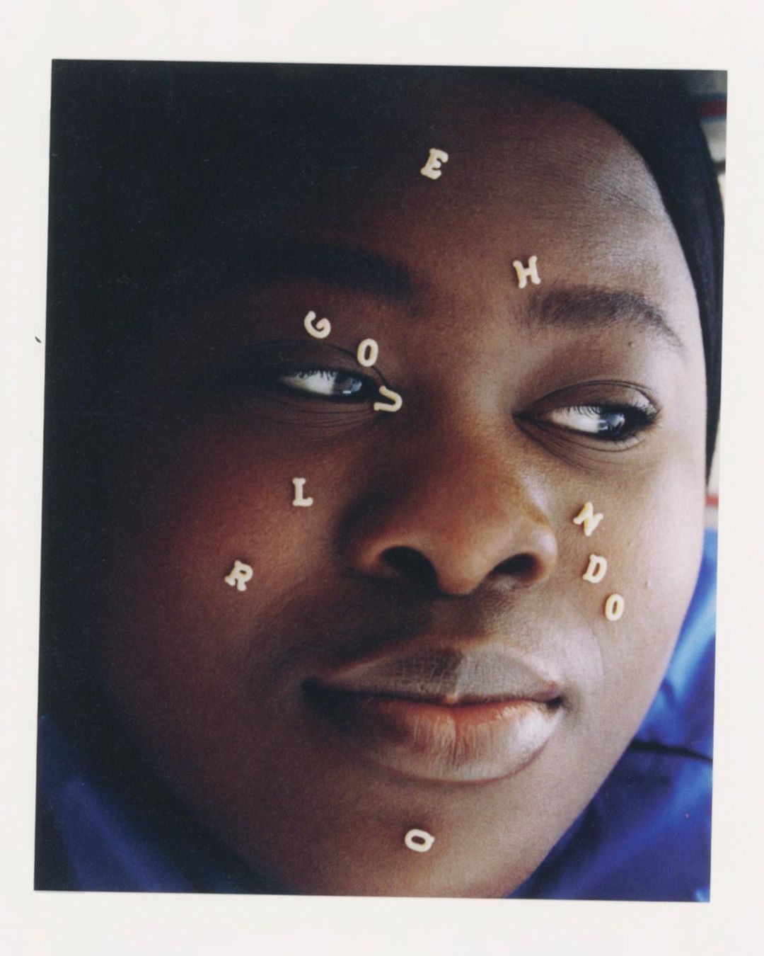 Close-up portrait of a Black woman with alphabet pasta letters on her face, eyes looking to the side, set against a plain background.