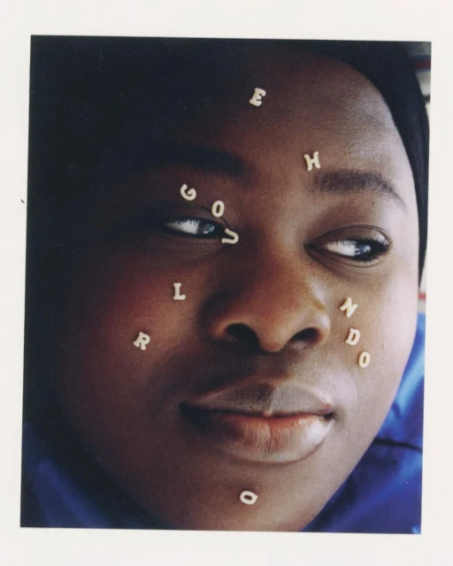 Close-up portrait of a Black woman with alphabet pasta letters on her face, eyes looking to the side, set against a plain background.