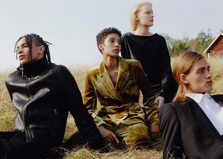 A group of four young adult fashion models are sitting on tall grass in an open field, posing and wearing stylish jackets and suits.