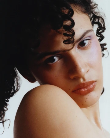 Close-up portrait of a woman with dark curly hair, wearing purple eyeshadow, against a plain white background.