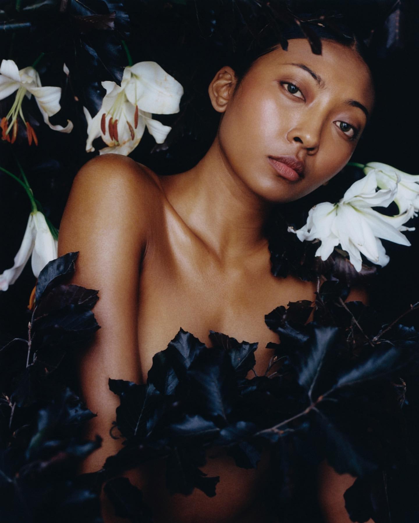 Portrait of a topless adult female with lily flowers and dark leaves in a dark environment, with a close-up framing and eye-level angle.