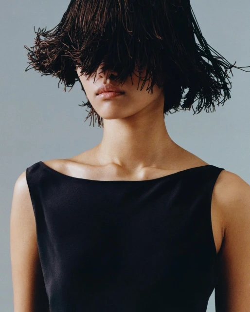 Headshot of a woman in a black dress with an elaborate dark brown fringe headpiece covering her eyes, set against a muted blue background.