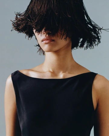 Headshot of a woman in a black dress with an elaborate dark brown fringe headpiece covering her eyes, set against a muted blue background.