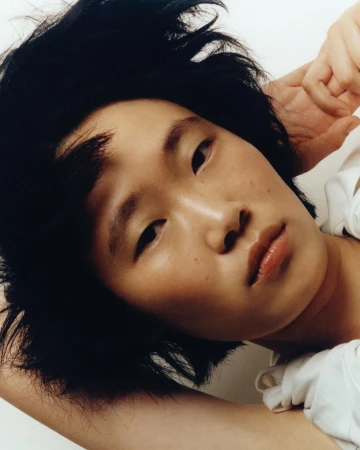 Close-up portrait of a young woman with short dark tousled hair and natural makeup posing against a plain background