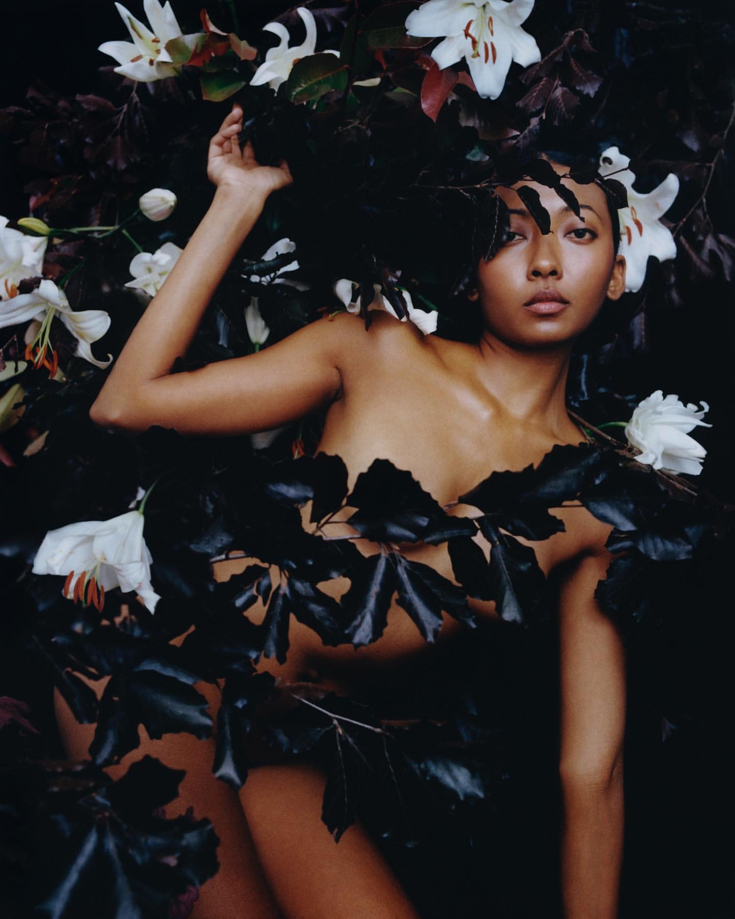 Woman poses partially covered in dark green leaves and white lilies in a studio shot. Portrait of model is eye-level and close-up.