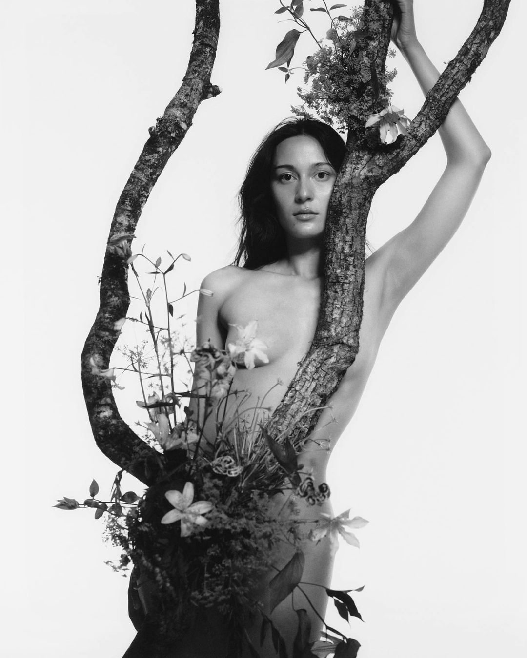 Monochrome portrait of a nude woman partially obscured by a blooming tree branch, creating an organic, ethereal visual effect.