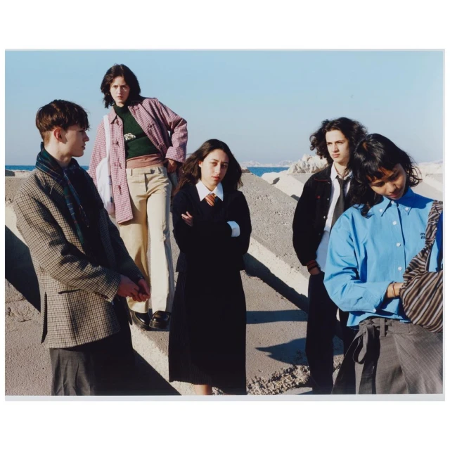 Group of five young adults standing on a concrete structure against a light blue sky, wearing casual clothing, with a distant sea view.