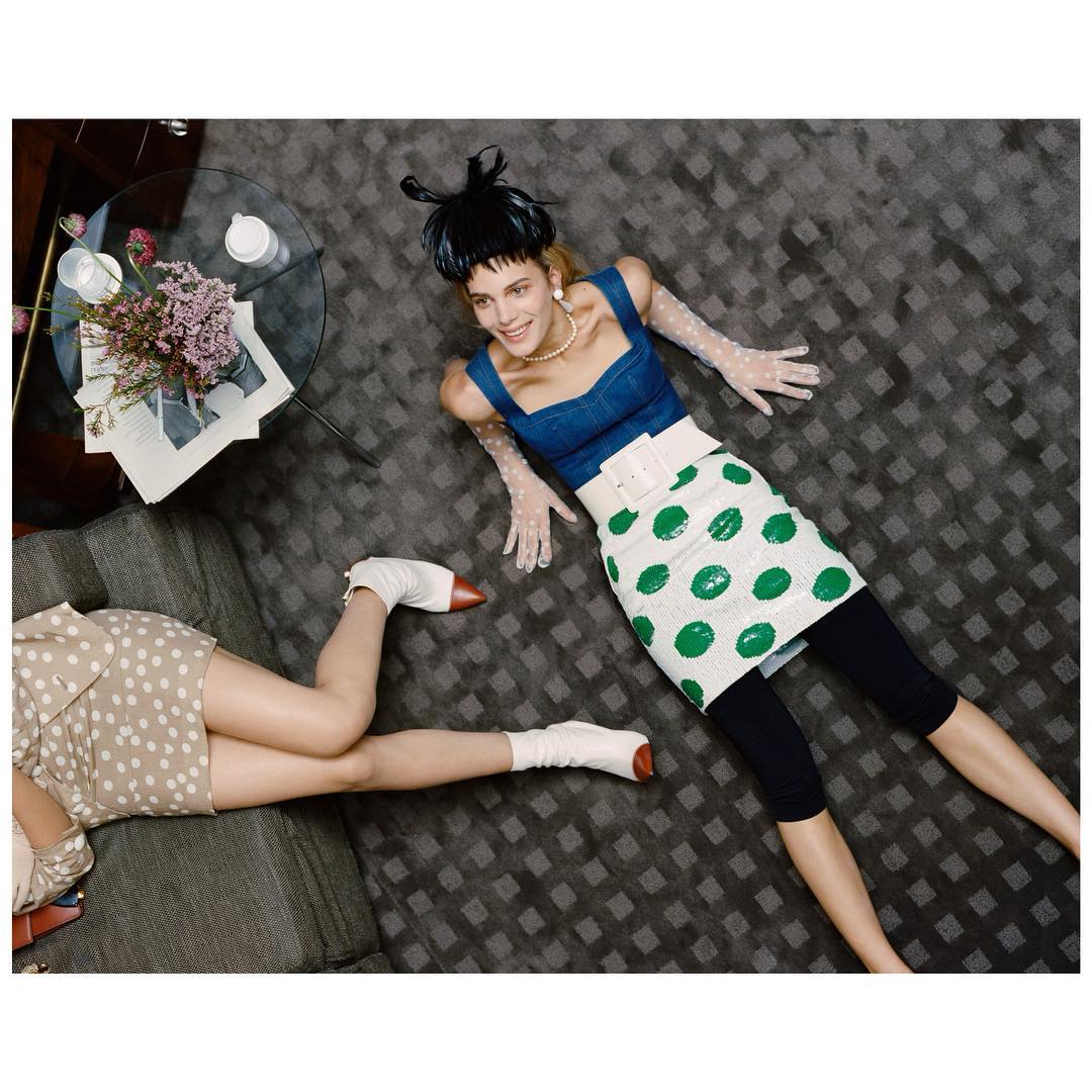 Overhead shot of a woman lying on a grey patterned rug wearing a blue top, white and green polka dot skirt, and white gloves, posing with another person.