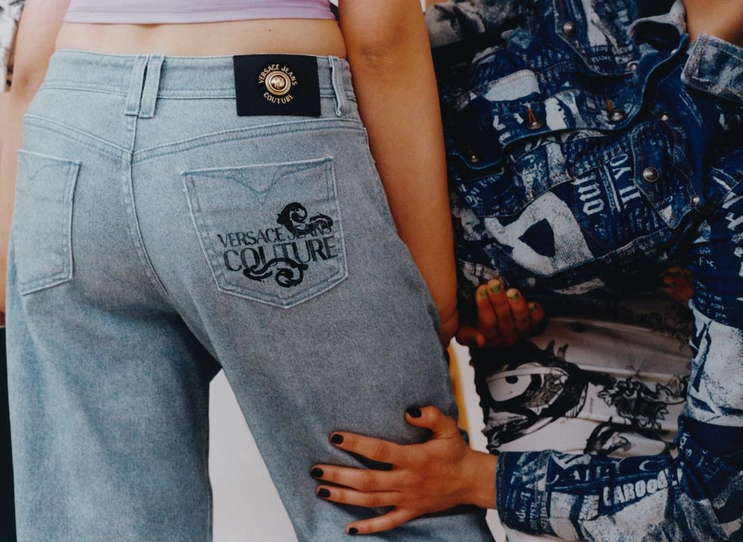 Close-up of woman in light-wash Versace jeans, hands on hips, next to another person in a patterned denim jacket. Fashion shot, studio lighting.