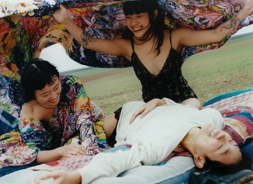 Three smiling Asian women relax outdoors on a blanket under a colorful floral textile, with a field and sky visible in the background.