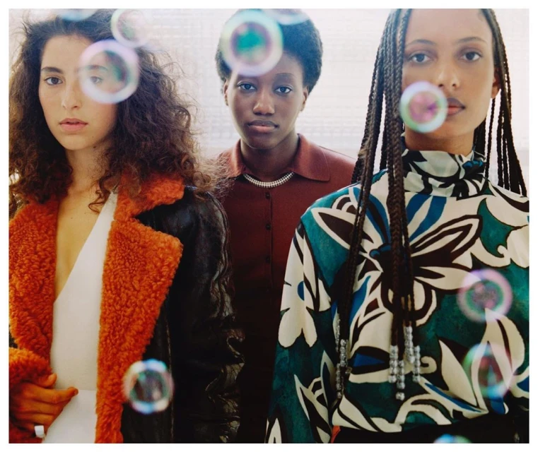 Studio shot features three women of diverse ethnicities in fashionable tops surrounded by bubbles, set against a neutral background.