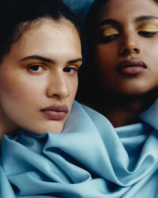Close-up shot of two women with yellow eyeshadow and mauve lipstick posing in front of a dark blue backdrop, wrapped in blue fabric.