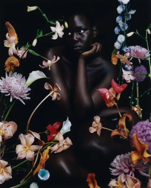 Striking portrait of a Black woman surrounded by an array of colorful flowers against a dark backdrop, creating a serene and beautiful image.