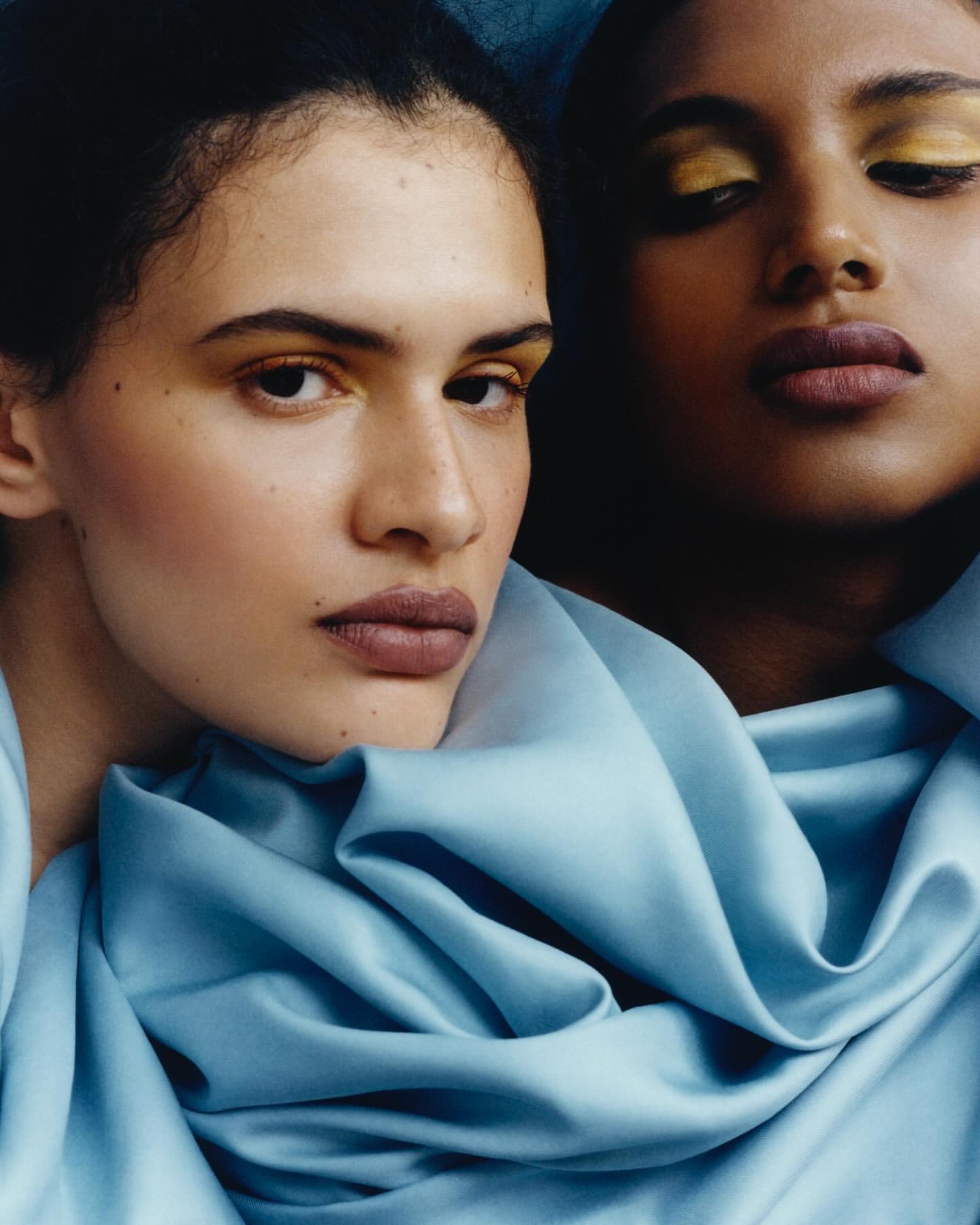 Close-up of two models with light and dark skin tones wearing gold eye makeup and matching pastel blue silky fabrics.