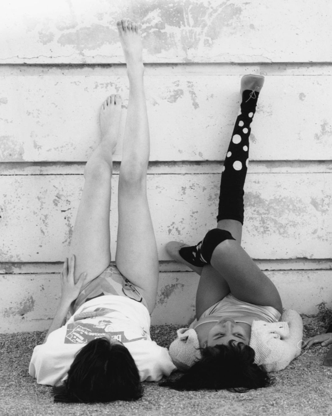 Black and white photo of two young women lying on the ground with their legs up against a textured wall, one wearing polka-dot leg warmers.