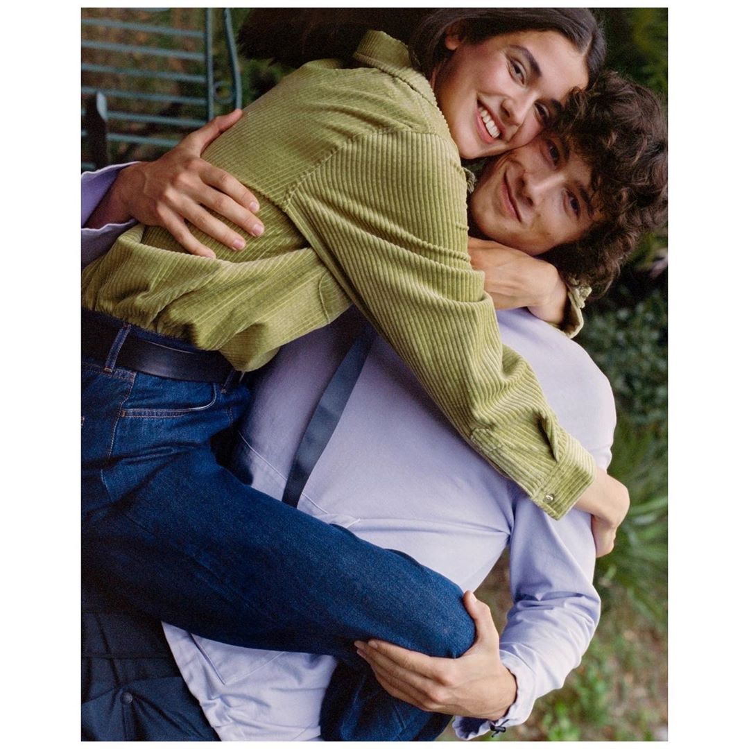 A smiling young couple in a close embrace outside. The woman wears a green corduroy jacket and the man wears a purple button-down shirt.