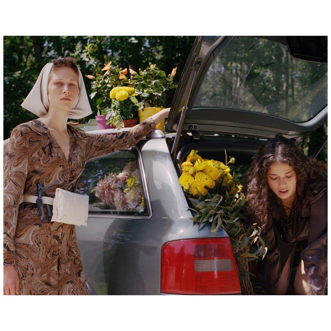Two women arrange colorful flowers in and around the trunk of a silver station wagon on a sunny day in a wooded setting.