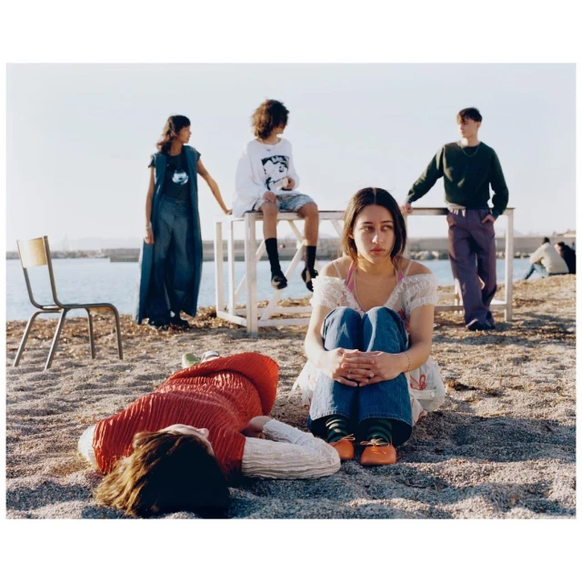 Group of five young people on a beach, one lying down, others sitting and standing near the water on a sunny day, wearing casual clothing.