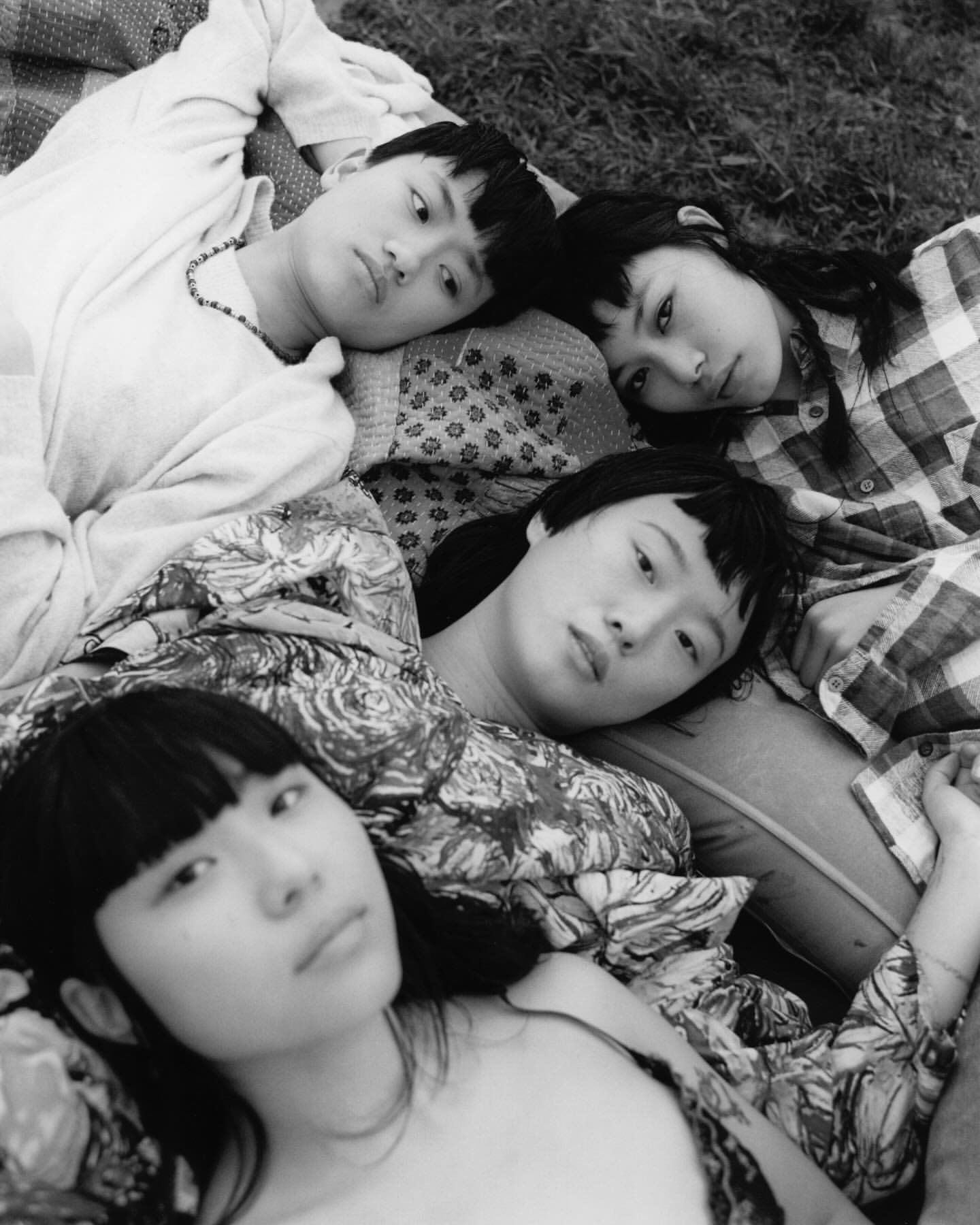 Black and white portrait of four young women lying on a patterned blanket in the grass, gazing directly at the camera with neutral expressions.